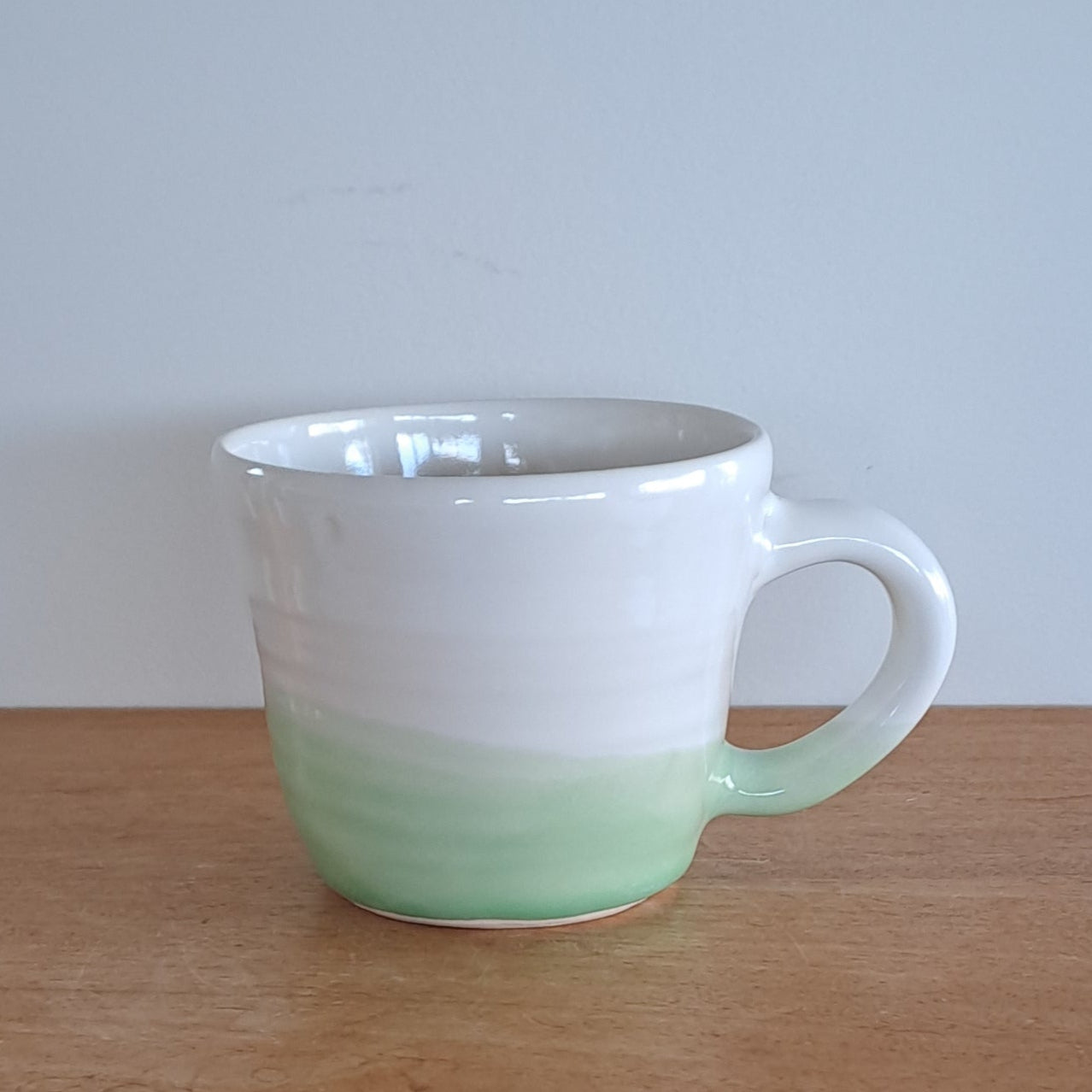 White and green cup on a wooden shelf against a white wall.
