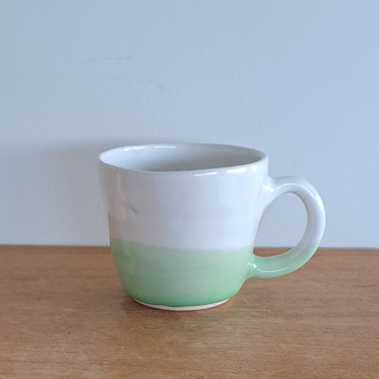 White mug with green base leaning against a wooden shelf.